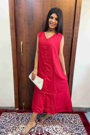 Woman wearing a sleeveless red cotton blend one-piece dress with front buttons, standing indoors on a patterned rug, holding a white clutch and wearing silver footwear."