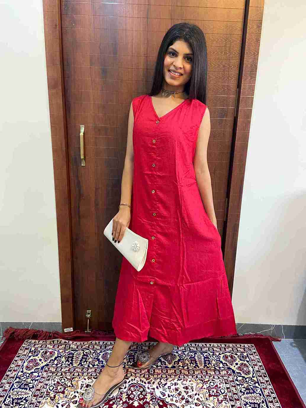 WhatsApp Image 2025-06-03 at 4.39.42 PM_11zon Woman wearing a sleeveless red cotton blend one-piece dress with front buttons, standing indoors on a patterned rug, holding a white clutch and wearing silver footwear."